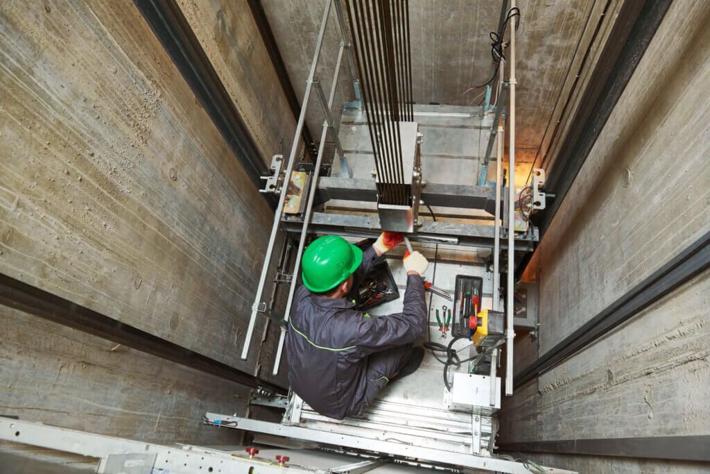 Elevator technician in green hardhat repairing cables inside elevator shaft.