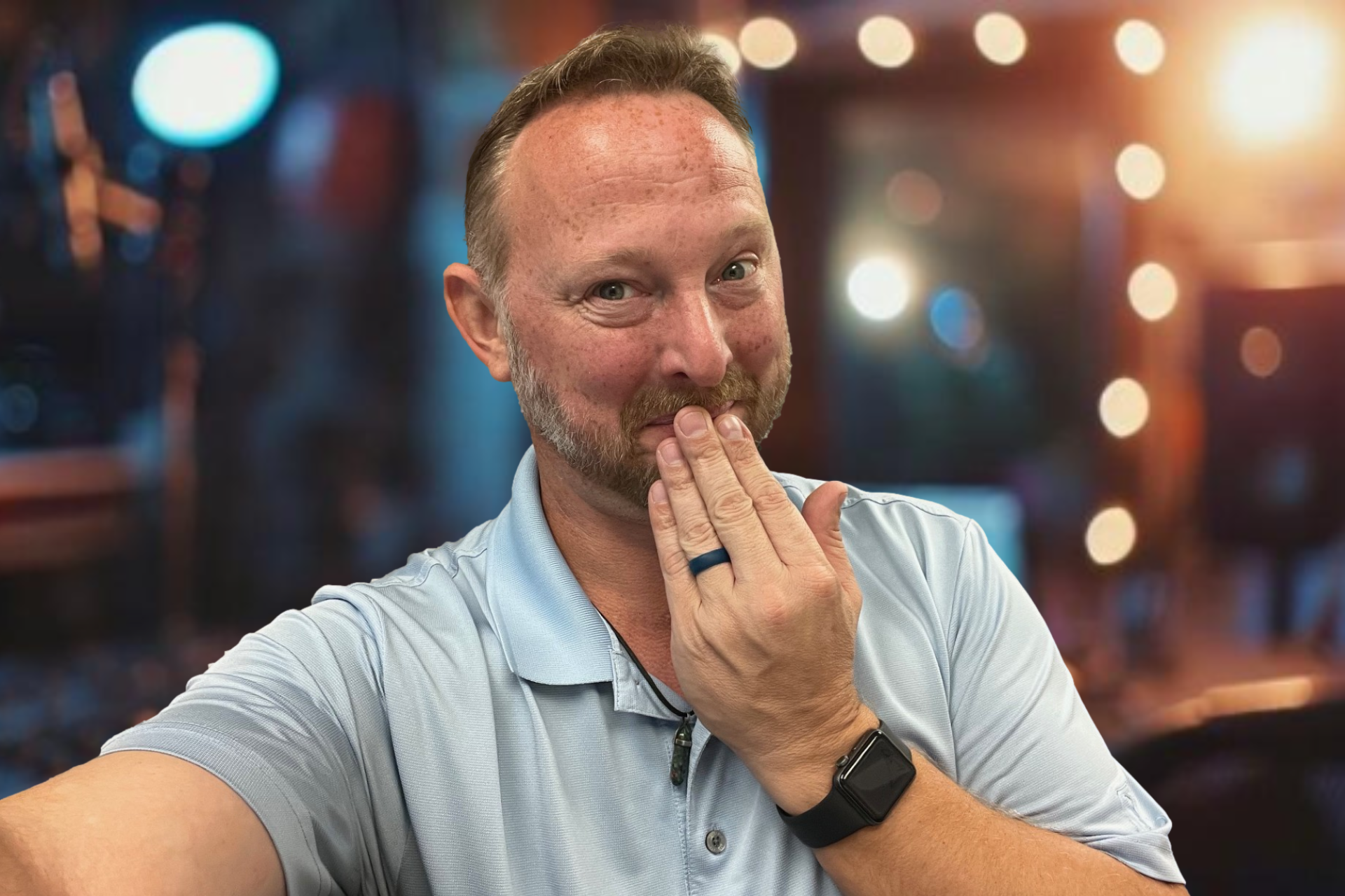 Man blowing a kiss, wearing a blue polo shirt and a watch, with a blurry outdoor background.