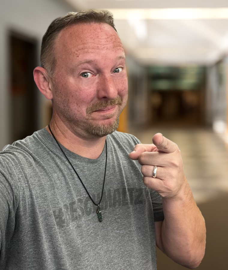 A man in a grey shirt is pointing at the camera.