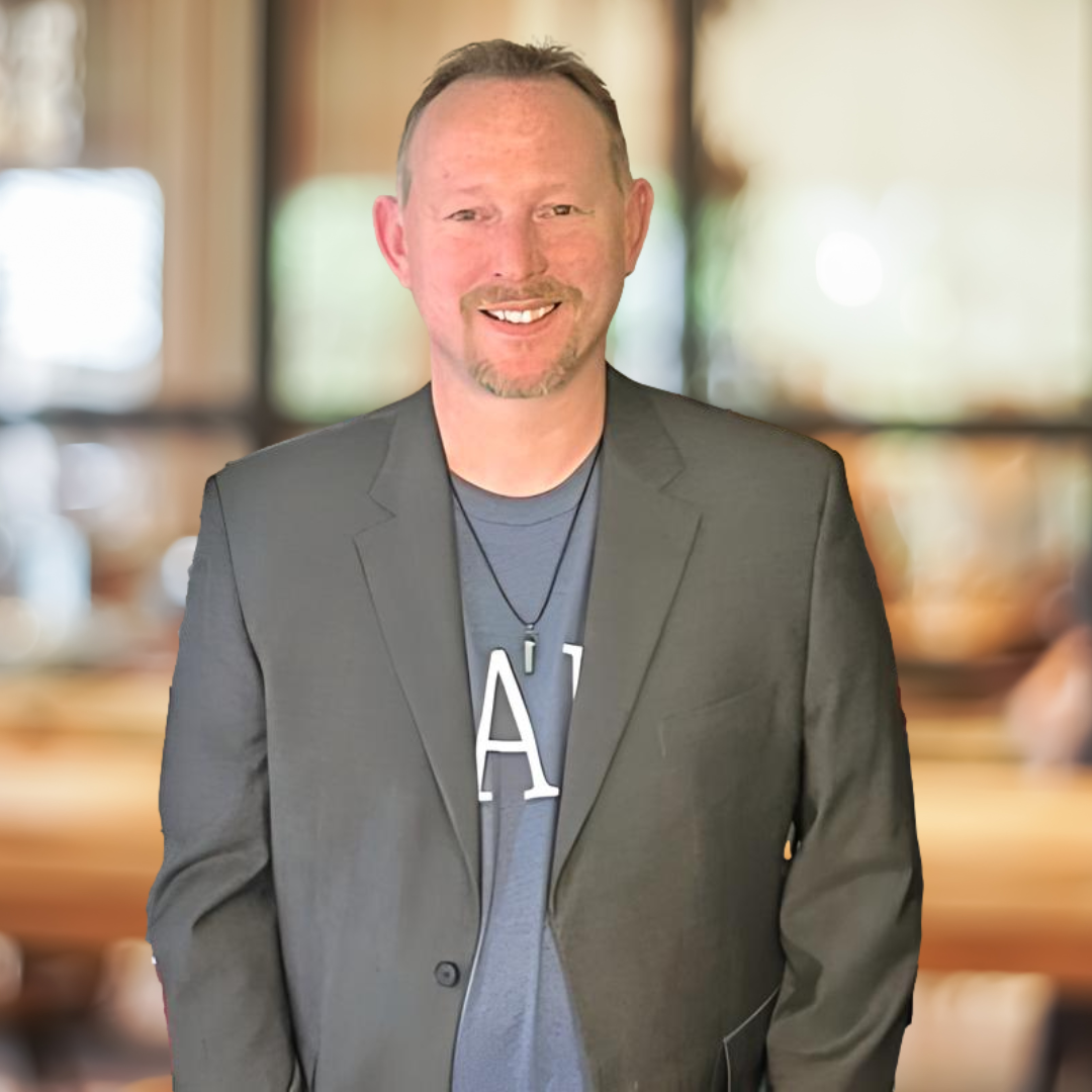 Tony Schmaltz in a suit and a blue shirt with the letter a on it is smiling for the camera.