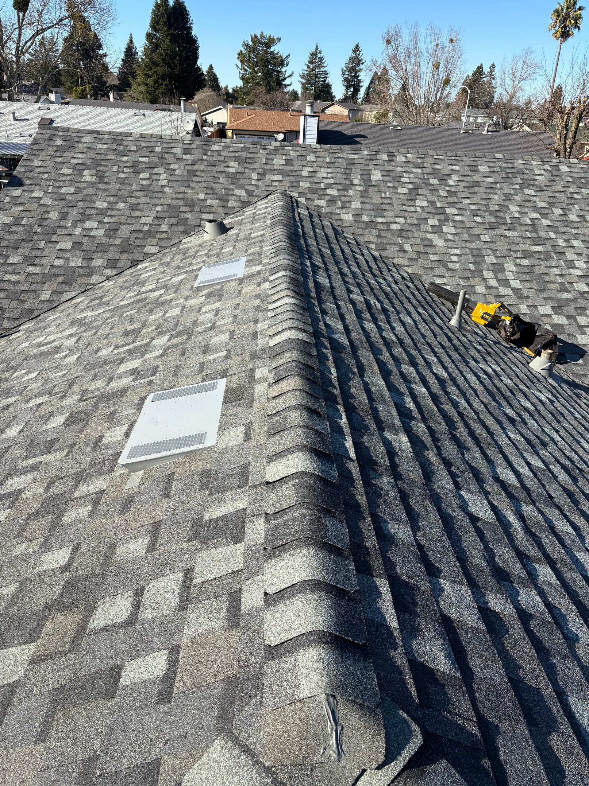 Gray asphalt shingle roof, angled view, with roof vents and distant buildings under a clear sky.