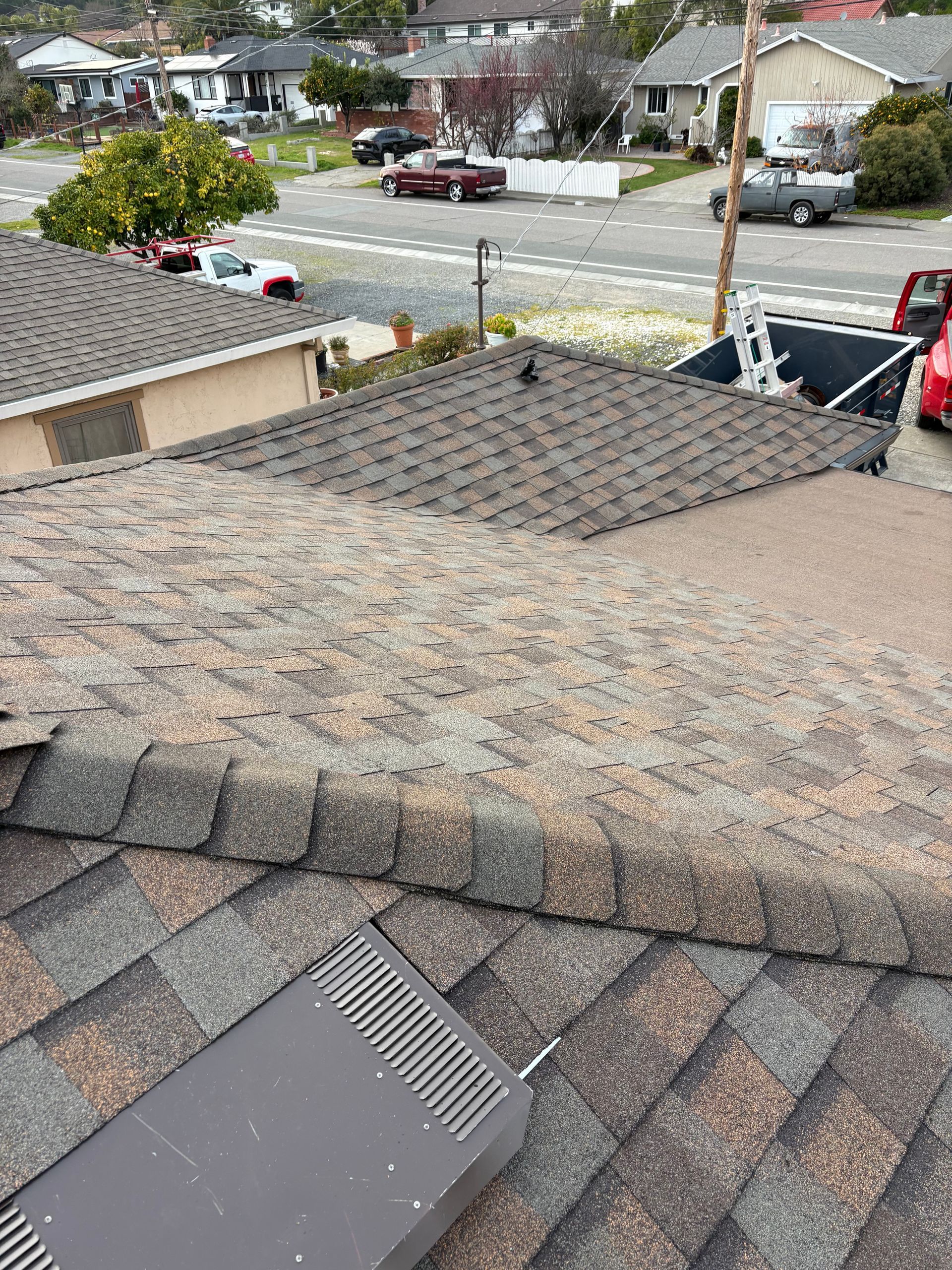 View of a roof with brown and gray shingles. A metal vent and neighborhood houses are in the background.