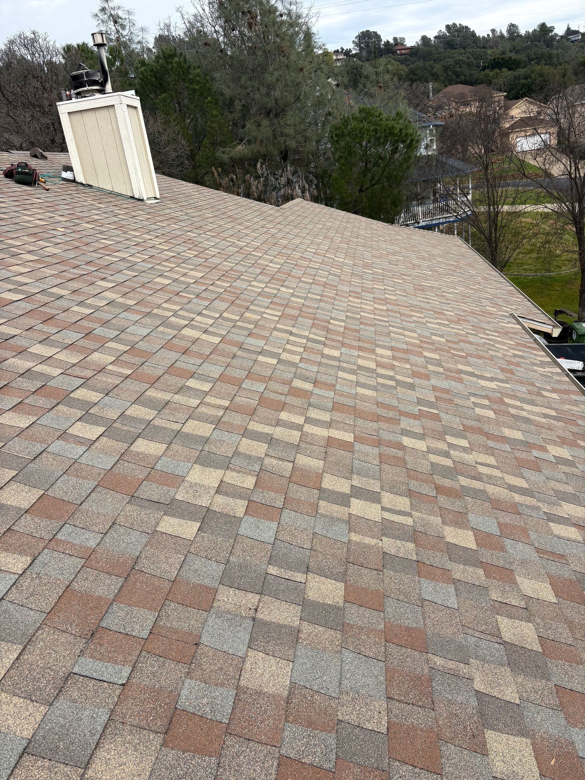 Close-up of a multi-colored asphalt shingle roof with a chimney and trees in the background.