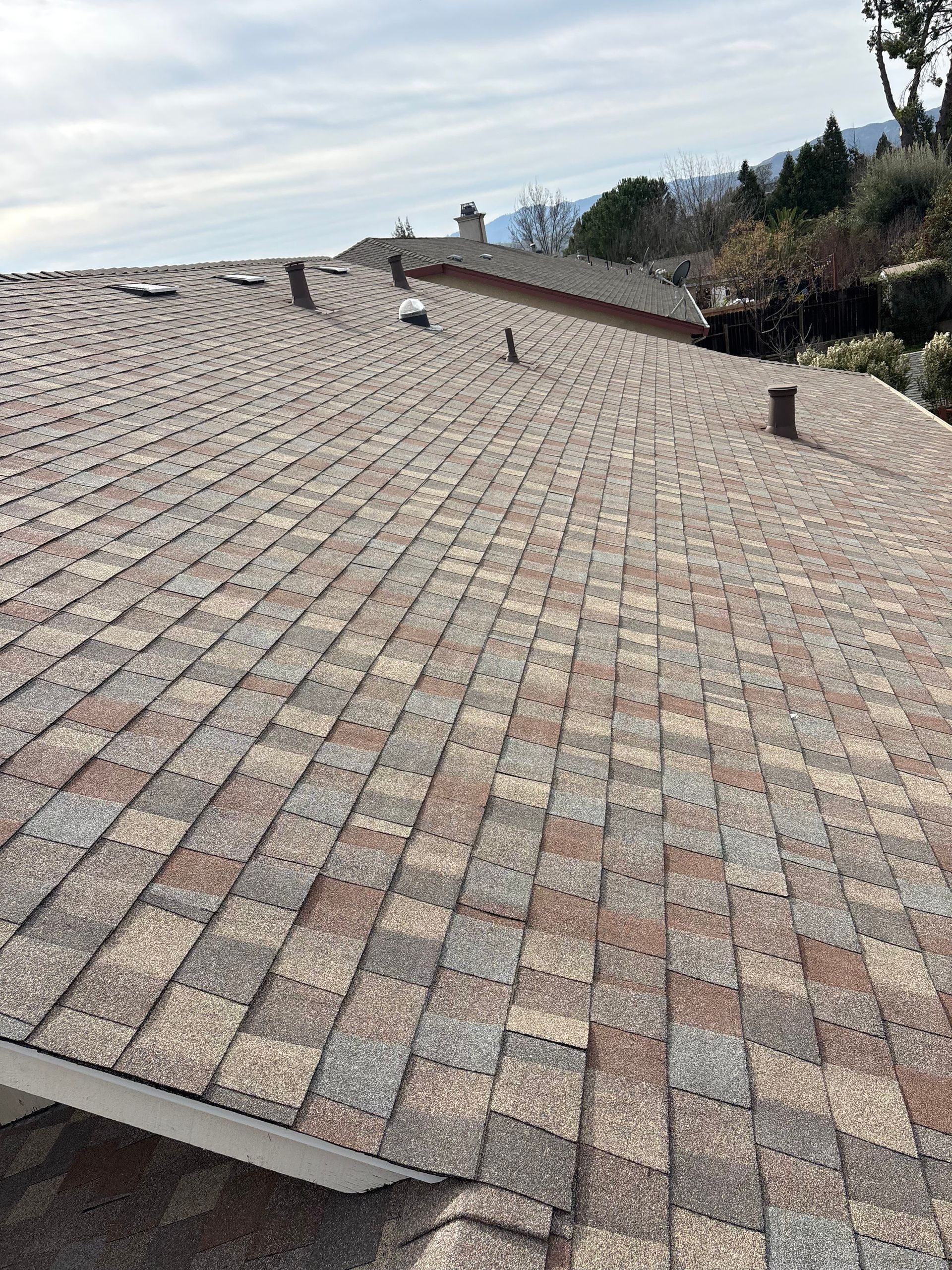 Brown and tan asphalt shingle roof on a house, with several vents and a slightly cloudy sky.