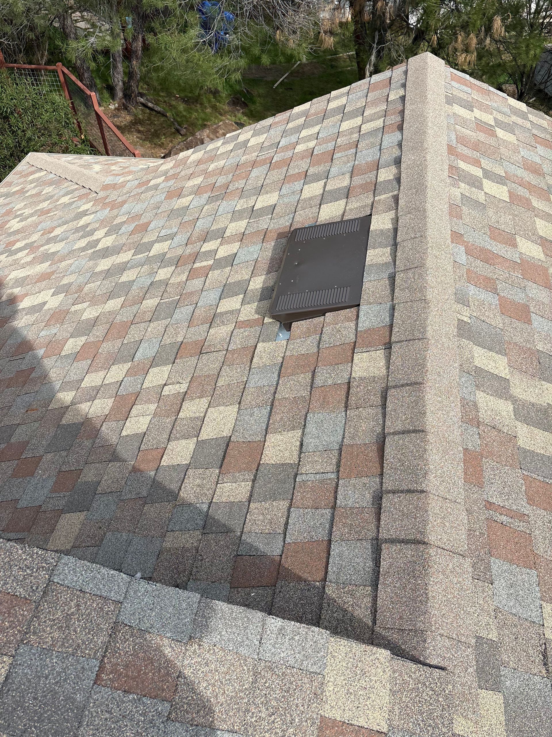 Close-up of a multi-colored shingle roof with a vent and a forest in the background.