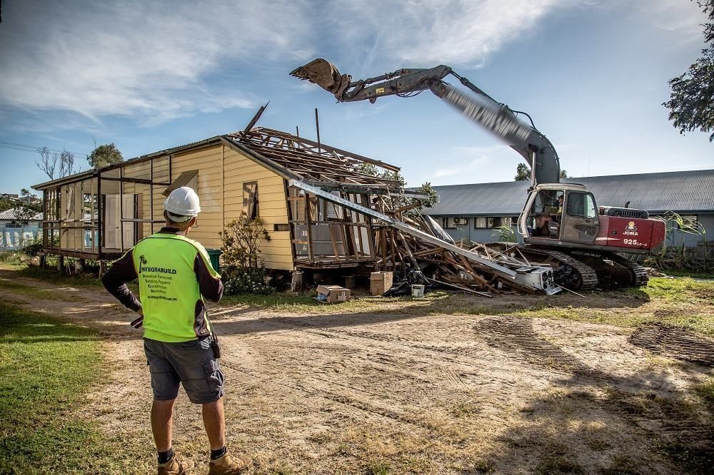 A Man Is Standing In Front Of A House Being Demolished By A Large Excavator — Integrabuild in Bowen, QLD