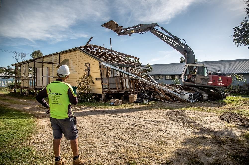 A Man Is Standing In Front Of A House Being Demolished By A Large Excavator — Integrabuild in Bowen, QLD
