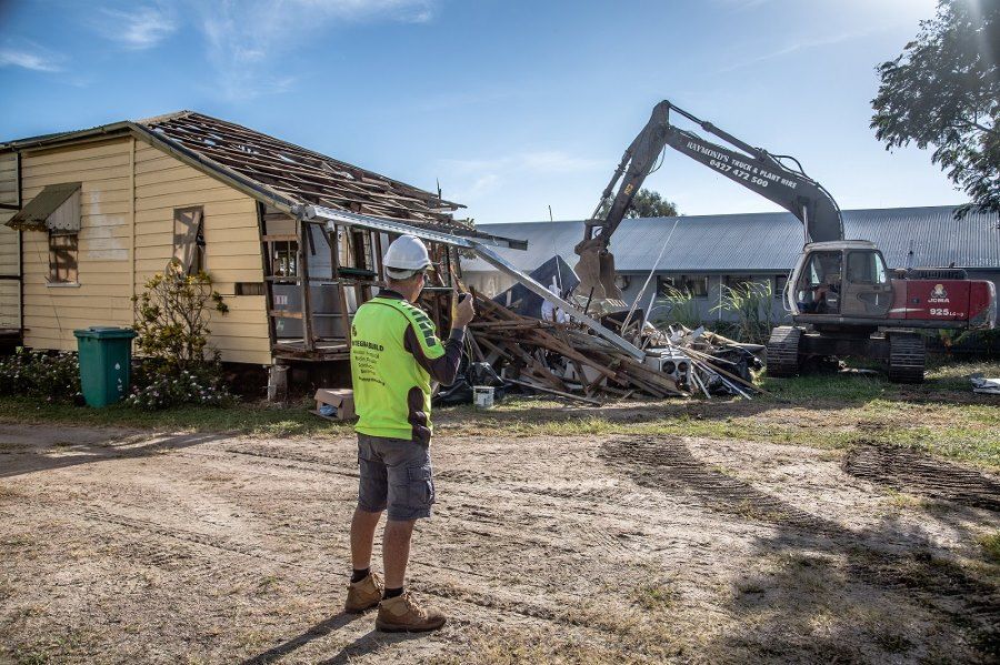 A Man Is Standing In Front Of A House Being Demolished By An Excavator — Integrabuild in Airlie Beach, QLD