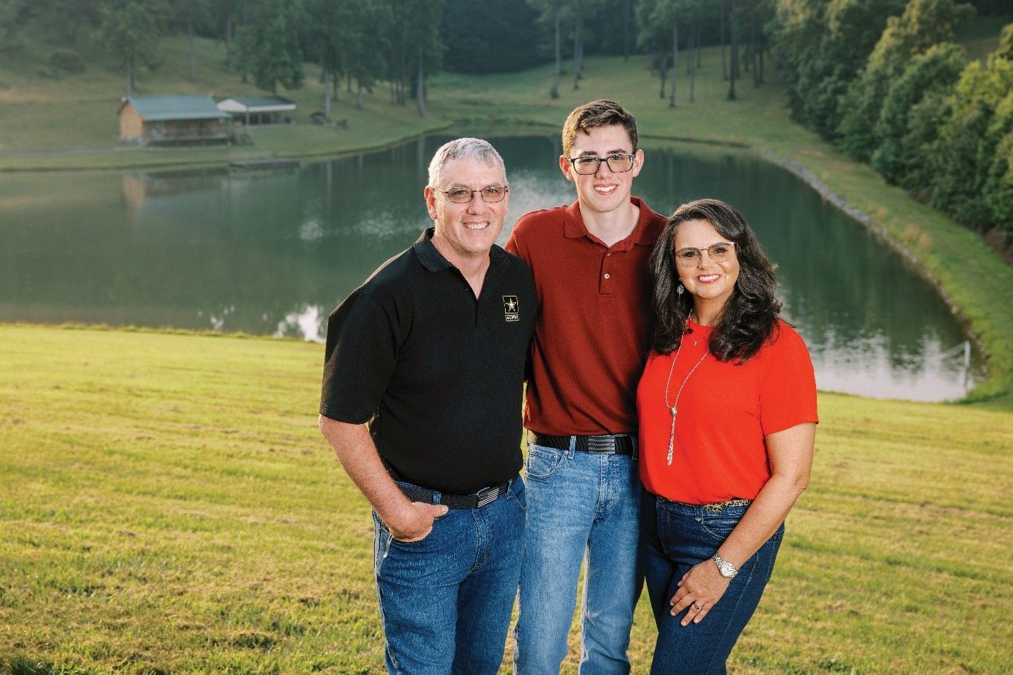 Family of three smiles outdoors near a pond, man in black shirt, son in red, woman in red.
