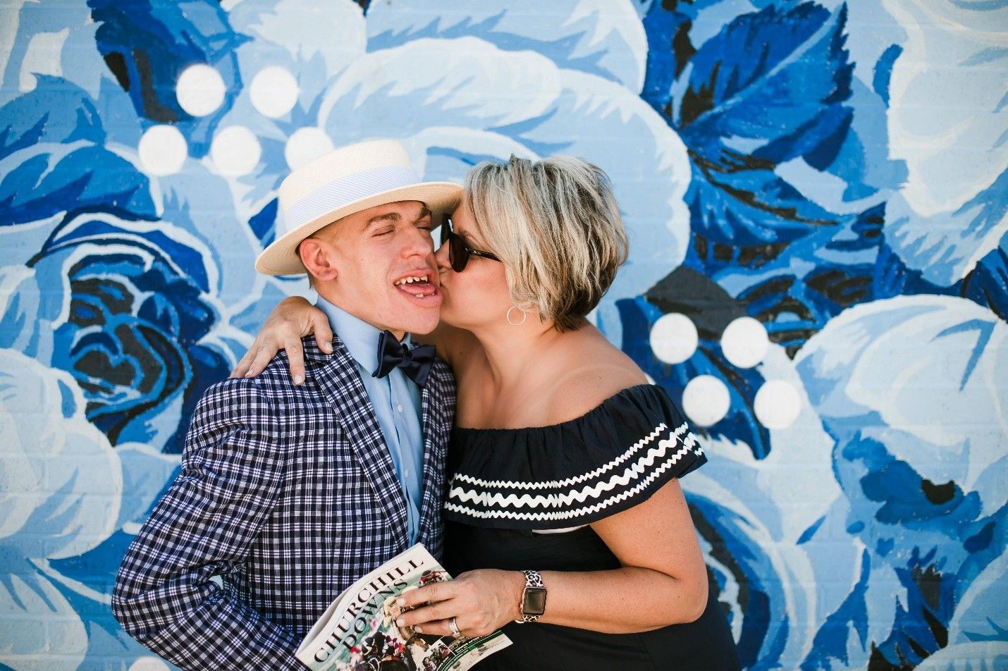Woman kissing a man wearing a hat and blazer in front of a blue floral mural.