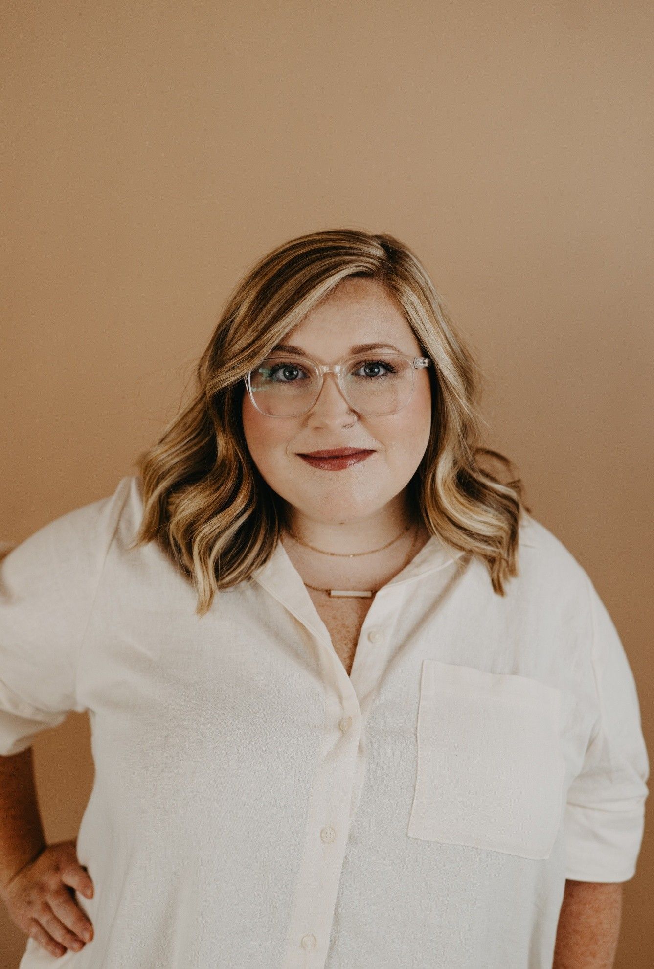 Woman with blonde hair and glasses in a light shirt, standing against a beige wall, smiling.