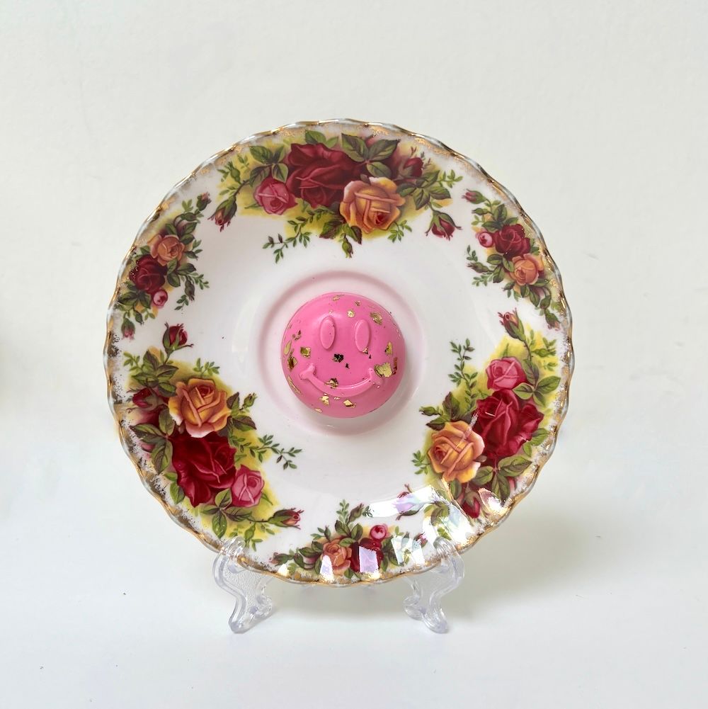 Een vintage schoteltje met bloemenmotief en gouden rand, waarop een roze smiley-bedeltje ligt