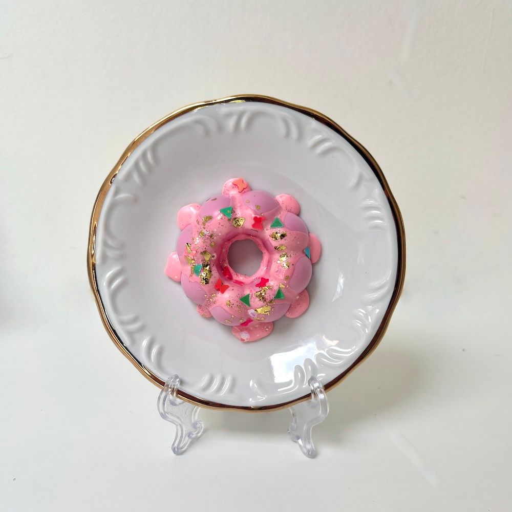 Een roze, bloemvormige donut met gouden en accenten, rustend op een klein wit schoteltje met een goudkleurige rand.