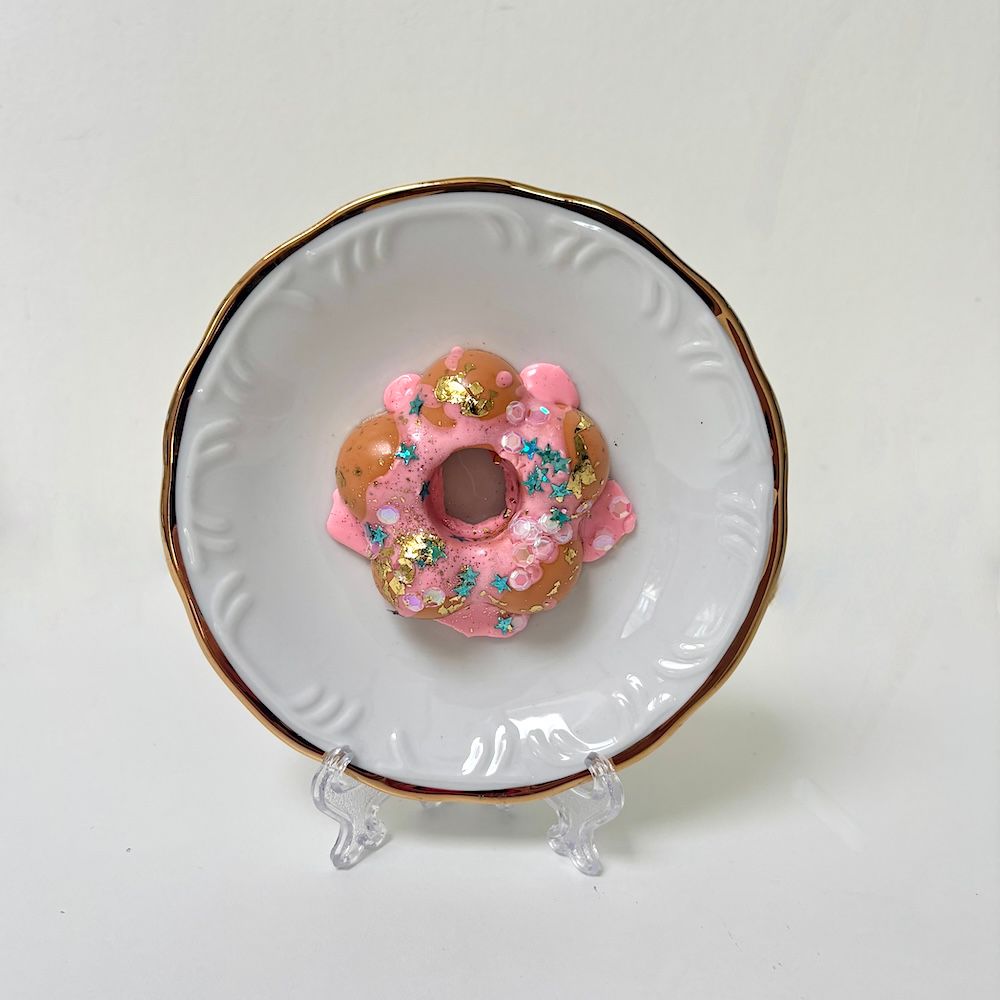 Een donutvormige sculptuur met roze glazuur, bladgoud en blauwe hagelslag, tentoongesteld op een klein, wit, sierlijk bordje.