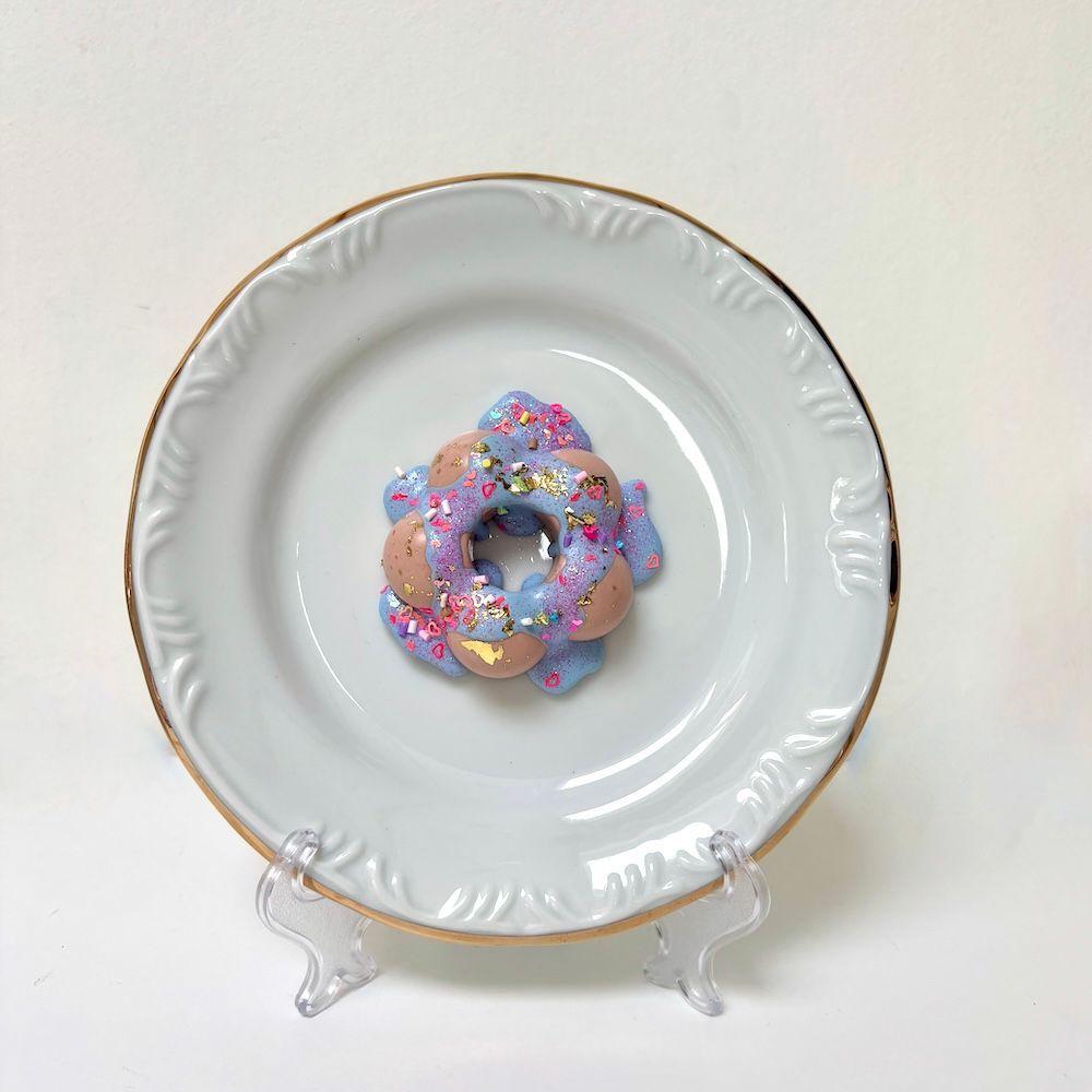 Een glinsterend, lavendel- en perzikkleurig donutvormig ornamentje ligt in het midden van een wit bord met een gouden rand.