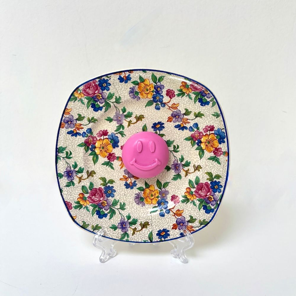 Een vintage vierkant bord met bloemenpatroon op een transparante standaard, met in het midden een felroze smiley.
