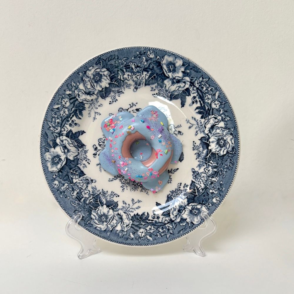 Een donut met blauwe glazuur en kleurrijke hagelslag ligt op een decoratief, blauw-wit keramisch bord met bloemenpatroon.