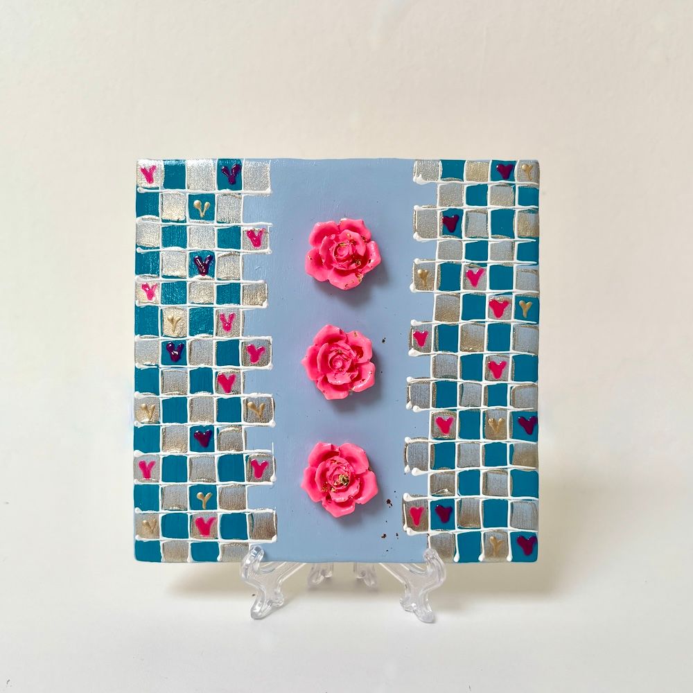 Een lichtblauwe tegel met drie roze rozen in het midden en mozaïekranden van zilveren en turquoise tegels aan de zijkanten.