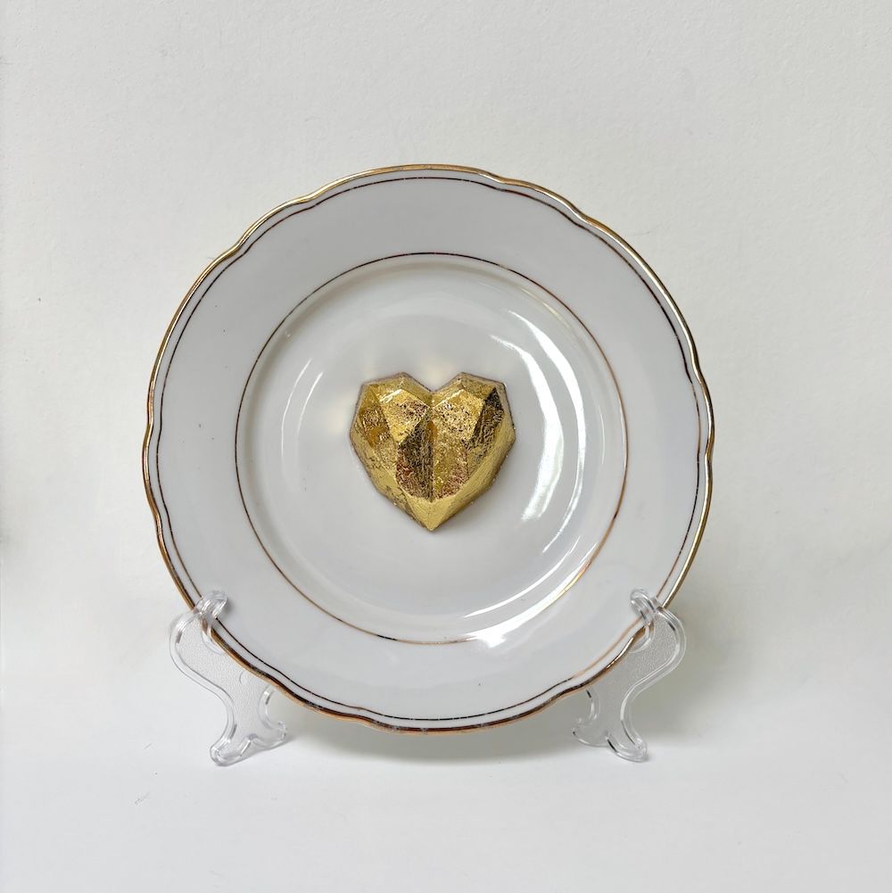 Een wit  bord met een gouden rand staat op een standaard met in het midden een gefacetteerd gouden hartvormig element.