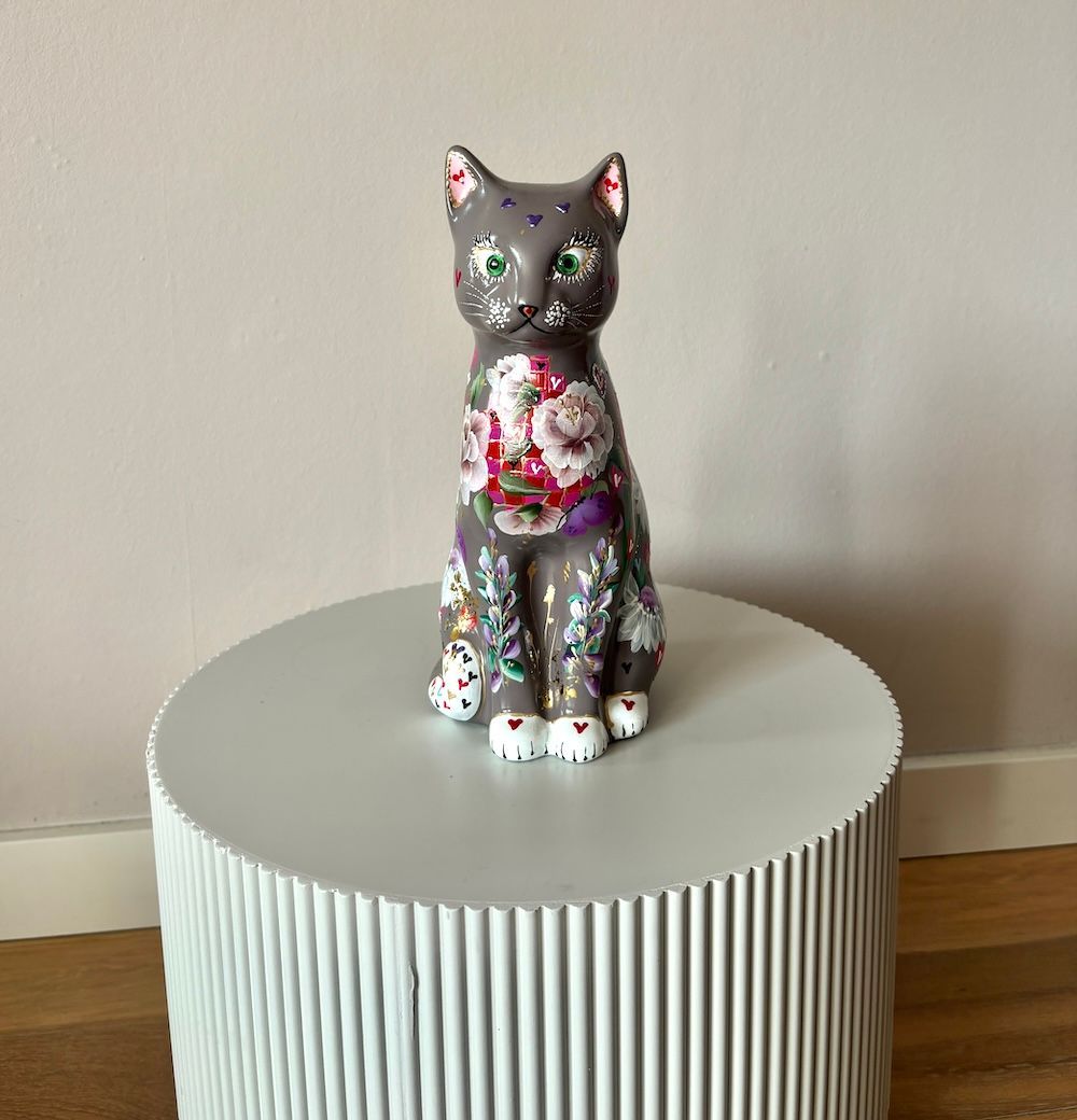 Een grijs, met bloemen beschilderd kattenbeeld staat op een, cilindrisch wit bijzettafeltje tegen een lichtgekleurde muur.