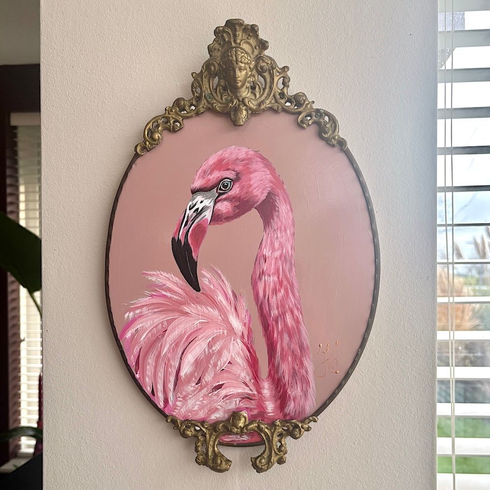 Een schilderij van een roze flamingo in een sierlijke, ovale gouden lijst aan een lichte muur.