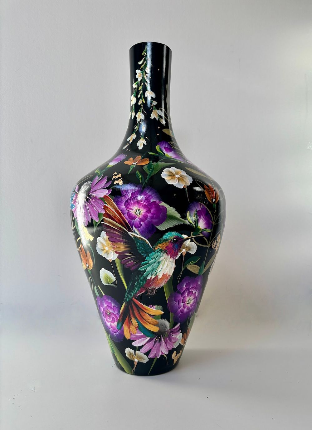 Een zwarte glazen vaas versierd met felpaarse bloemen, witte bloesems en een kleurrijke kolibrie.
