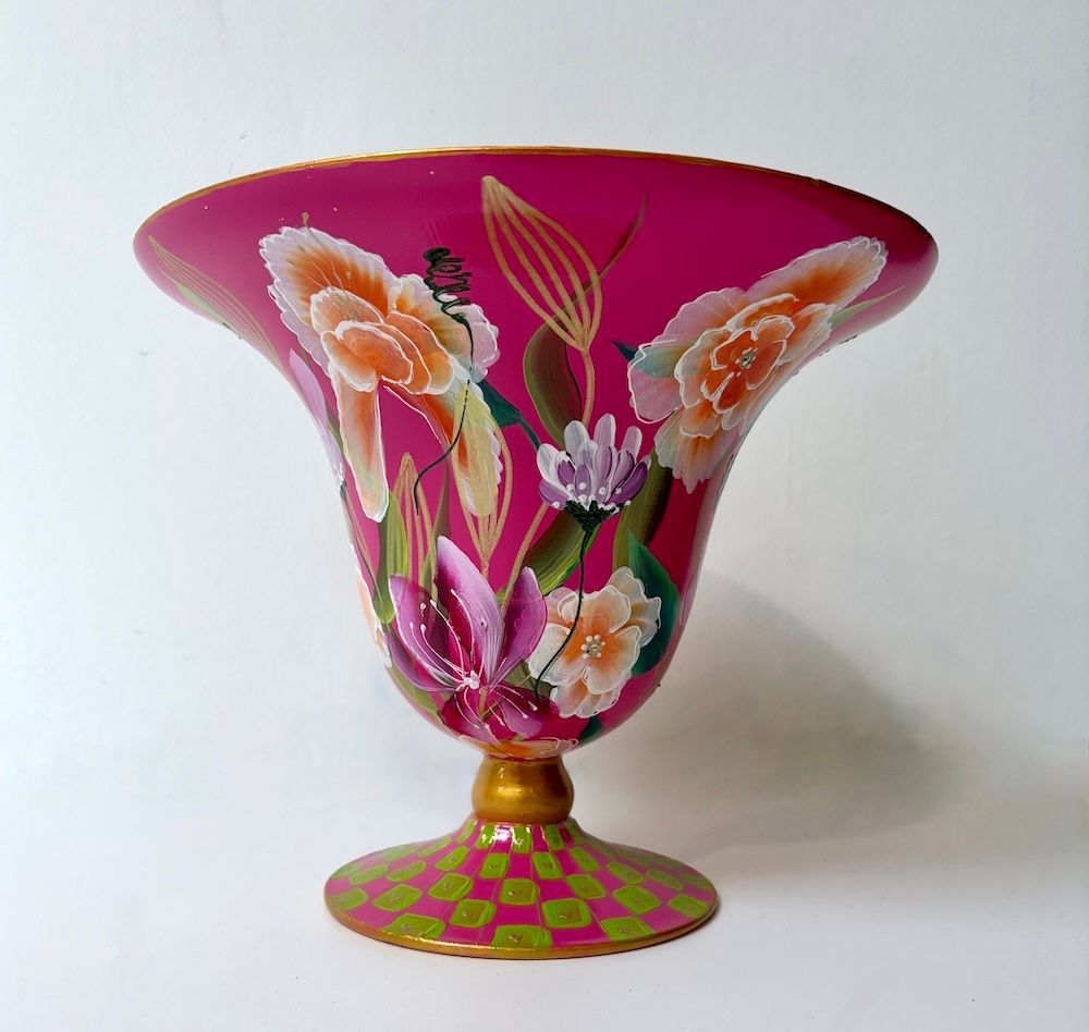 Een fuchsia glazen vaas op een voetstuk met beschilderde bloemmotieven, een gouden steel en een groen-roze geblokte voet.