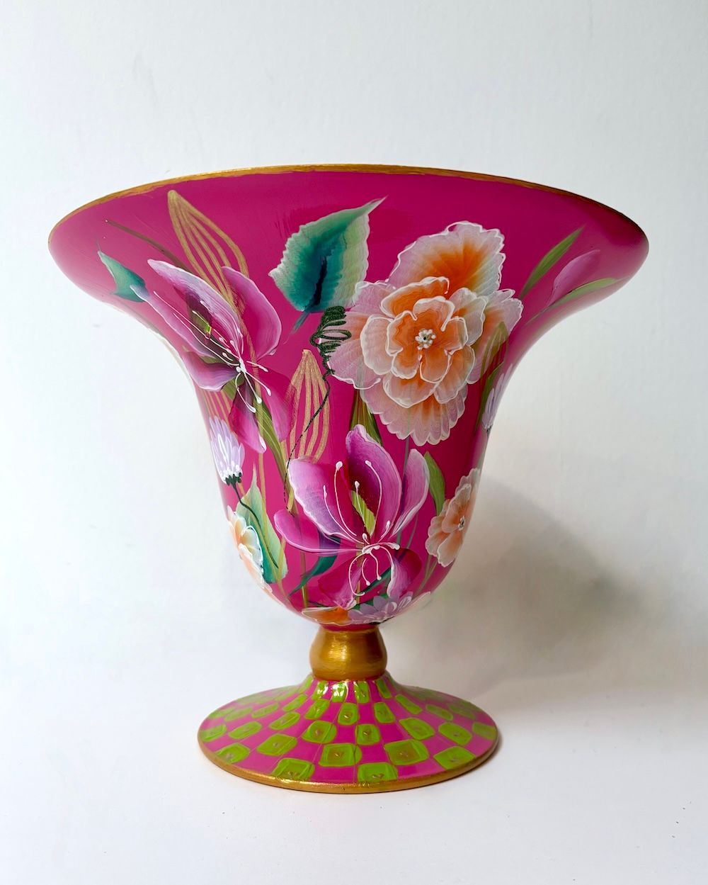 Een fuchsia glazen vaas op een voetstuk met beschilderde bloemmotieven, een gouden steel en een groen-roze geblokte voet.