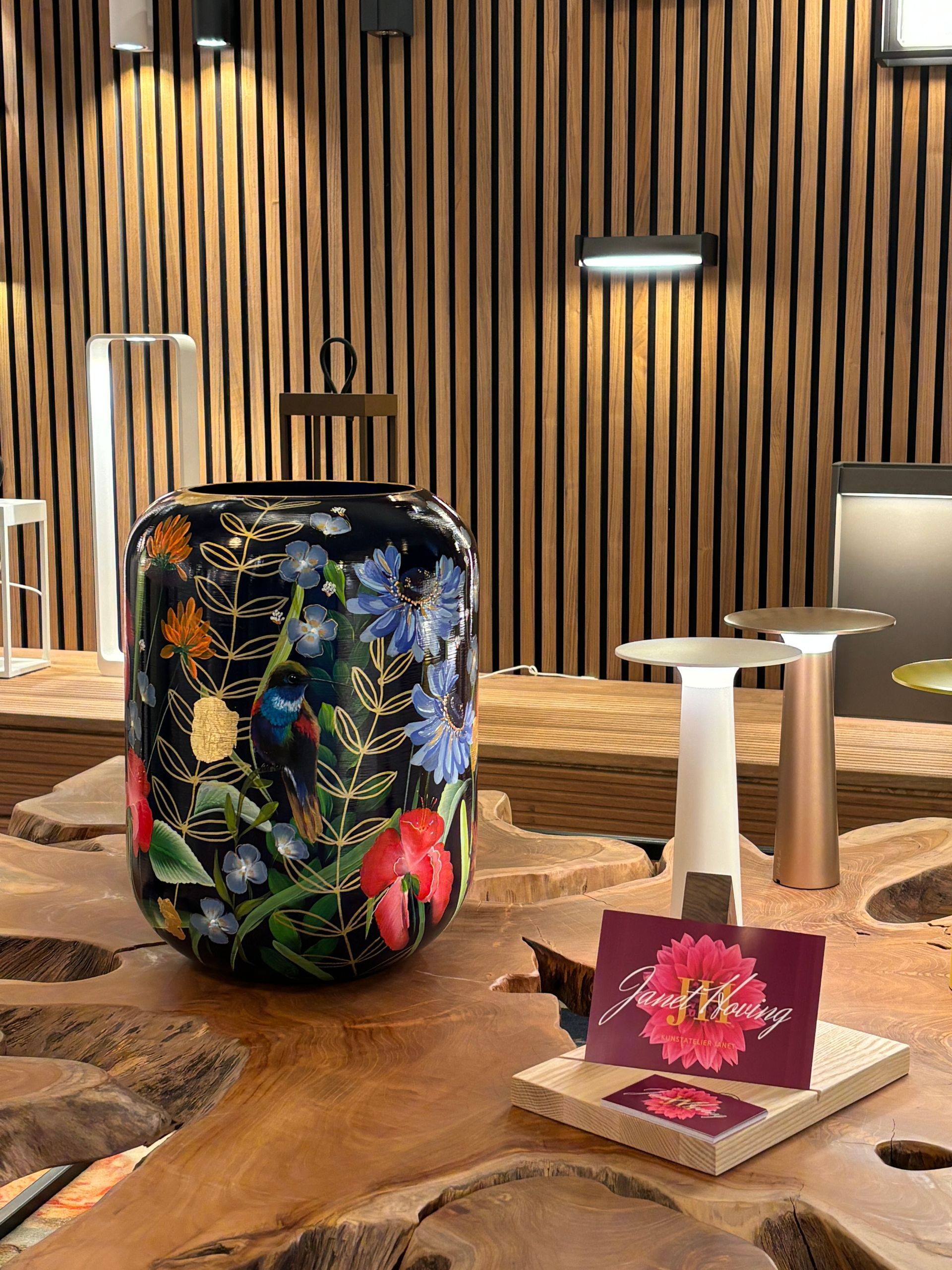 Een donker blauwe geschilderde cilinder vaas met bloemen staat op een houten tafel en een presentatiekaartje met een bloem.