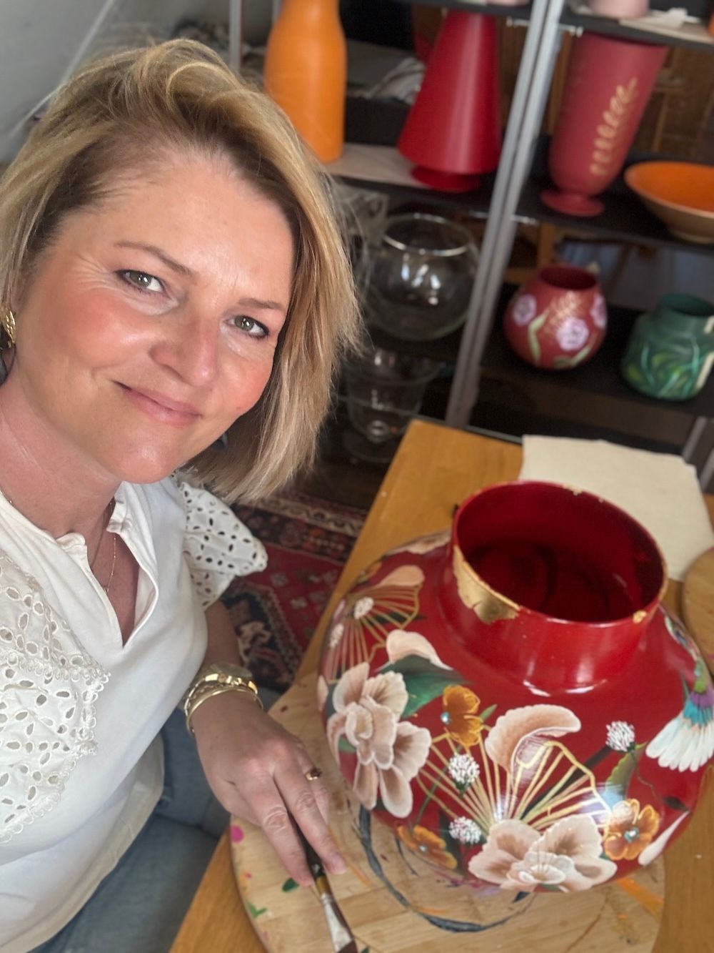 Een kunstenares is bezig met het beschilderen van een rode vaas met bloemenmotieven in een atelier vol decoratief glaswerk.