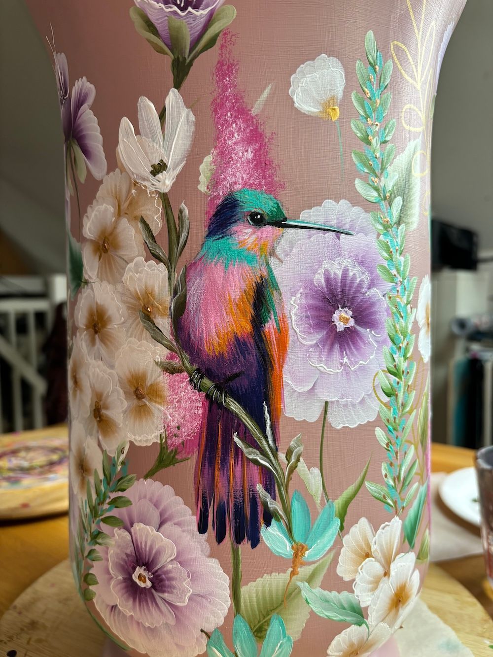 Een handbeschilderde vaas met een levendige kolibrie zit tussen roze, paarse en witte bloemen in een stoffig roze vaasje.