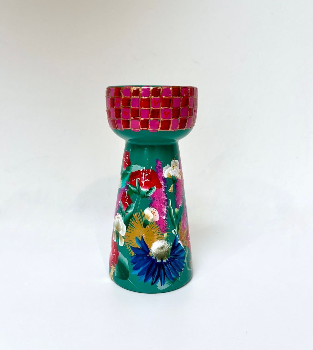 Een turquoise kaarsenhouder met een bovenrand van roze rode blokjes en handgeschilderde bloemmotieven op de voet