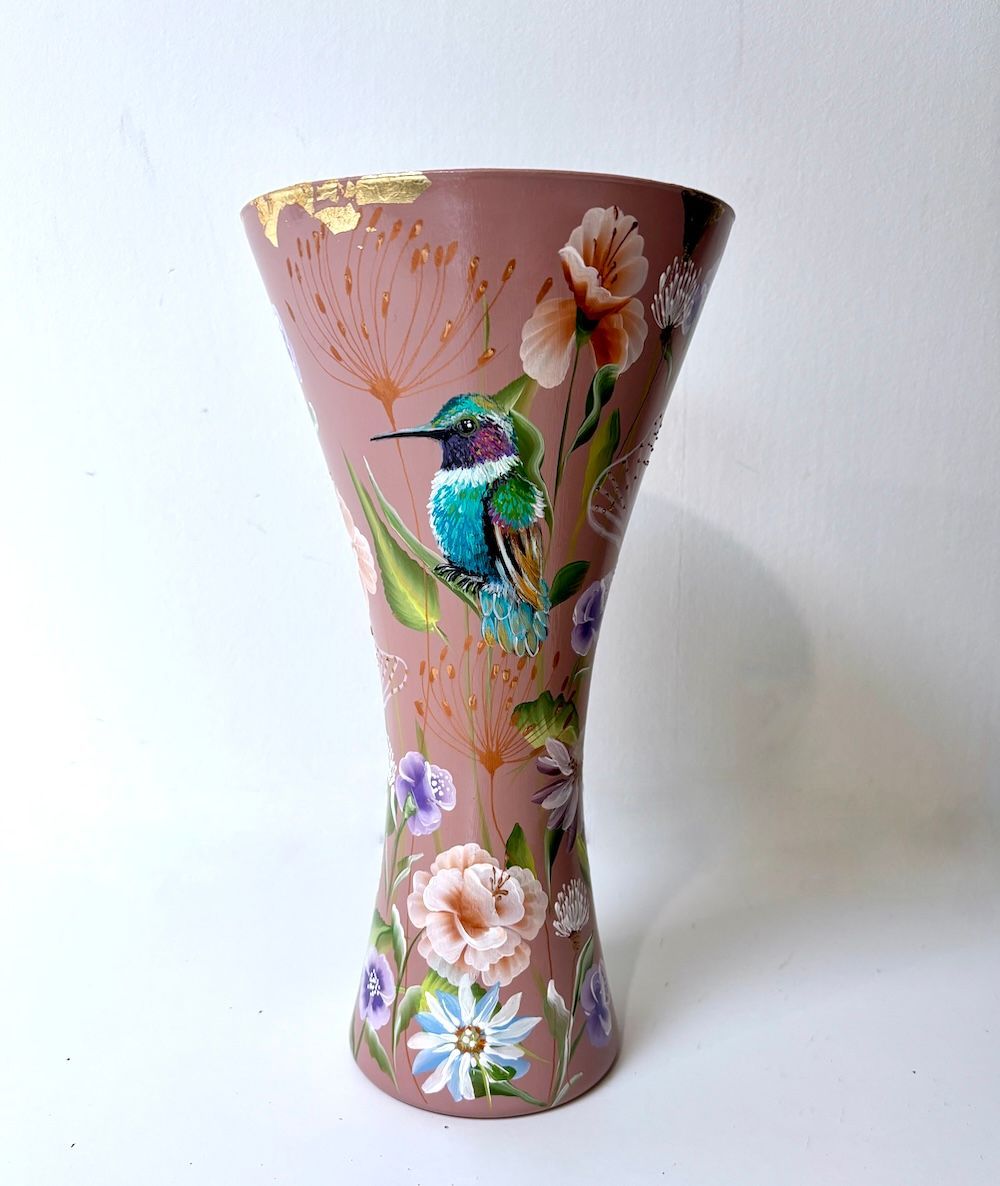 Een lichtbruine, zandlopervormige vaas, beschilderd met een kolibrie, diverse kleurrijke bloemen en accenten van bladgoud.