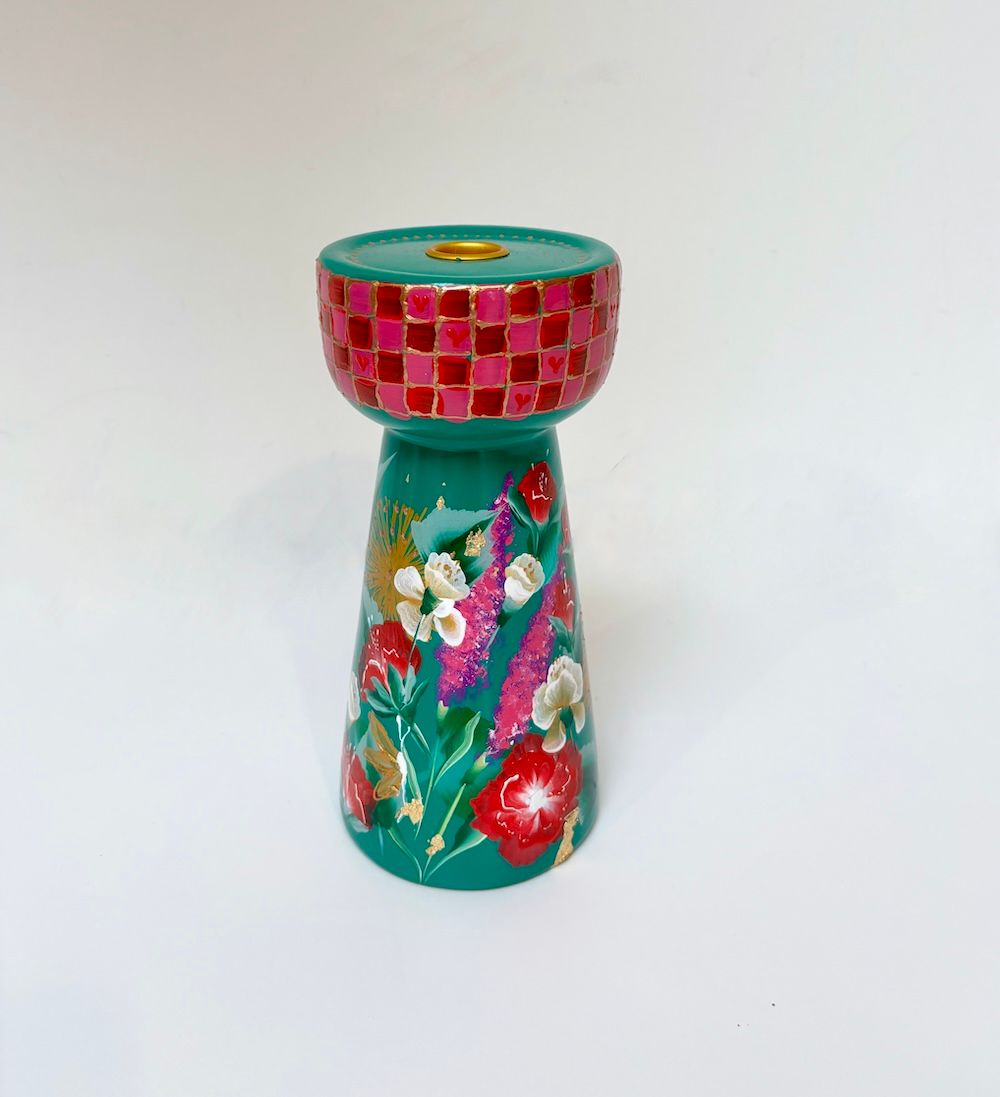 Een turquoise kaarsenhouder met een bovenrand van roze rode blokjes en handgeschilderde bloemmotieven op de voet