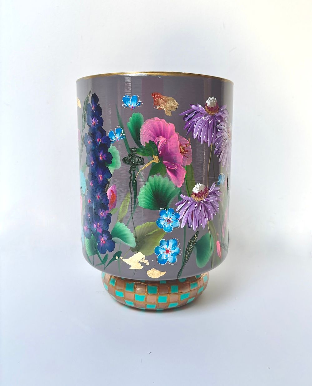 Een grijze vaas, beschilderd met  bloemen en groene bladeren, staat op een voetstuk met een patroon in turquoise en goud.