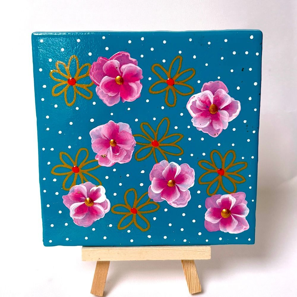 Klein beschilderd doek met roze bloemen en oranje bloemcontouren op een turquoise gestippelde achtergrond, op een schildersezel.