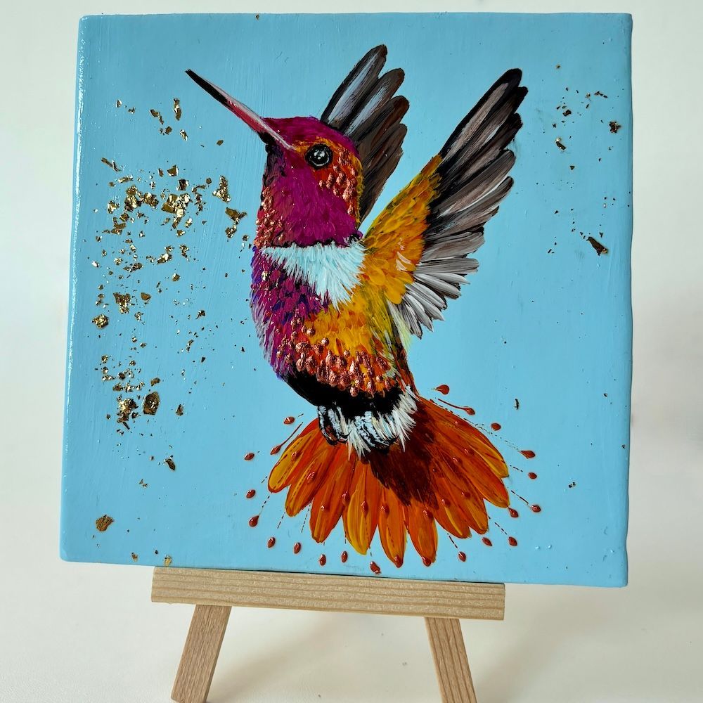 Kleurrijk schilderij van een kolibrie op een blauw canvas, tentoongesteld op een kleine houten schildersezel.