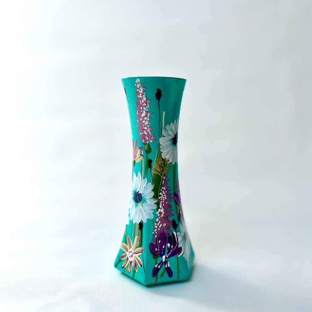 Een turquoise, zeshoekige vaas, handbeschilderd met witte en paarse bloemen, geplaatst tegen een effen witte achtergrond.