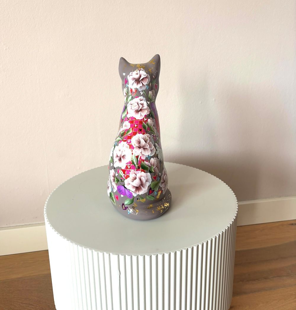 Een grijs, met bloemen beschilderd kattenbeeld staat op een, cilindrisch wit bijzettafeltje tegen een lichtgekleurde muur.
