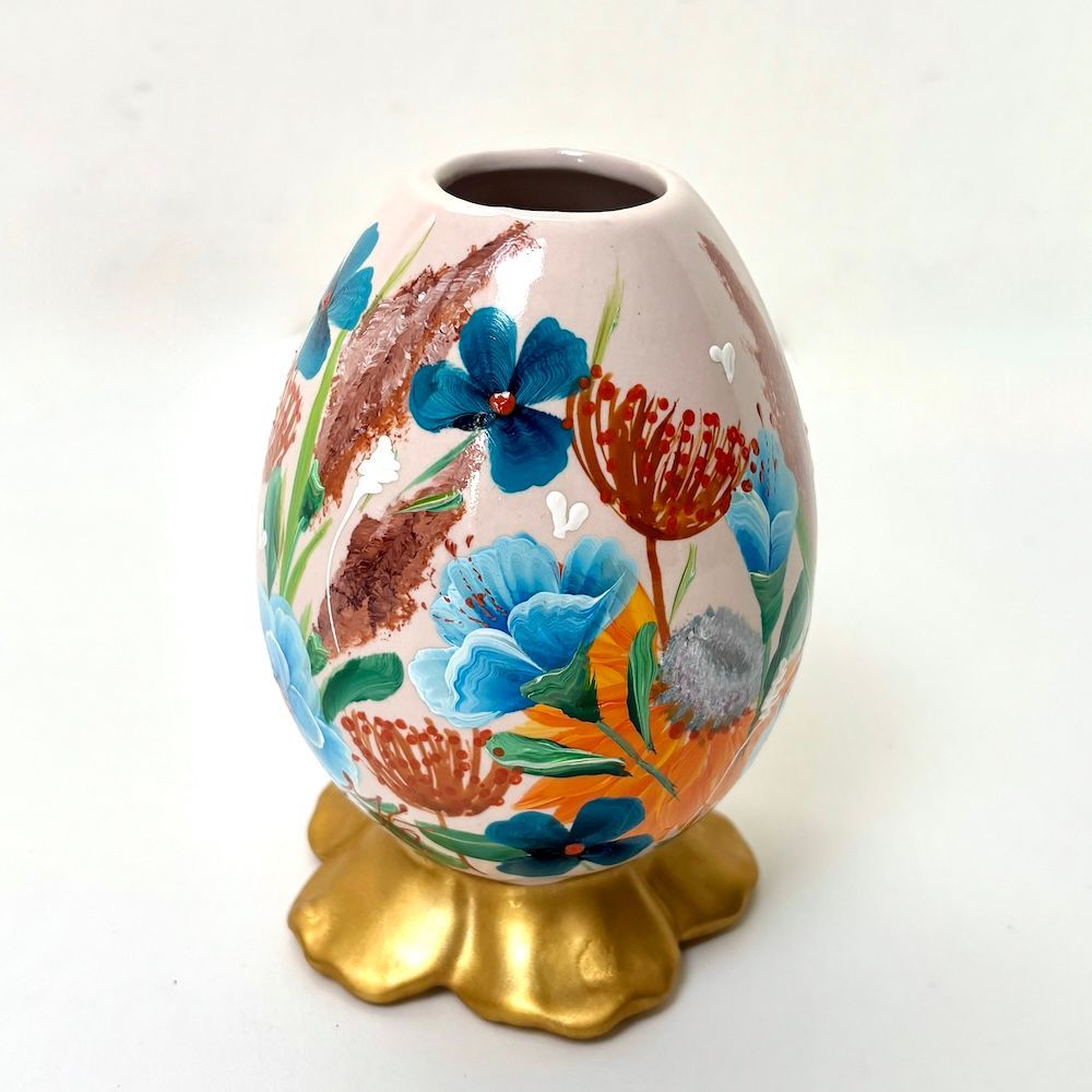 Een eivormige keramische vaas, beschilderd met blauwe en oranje bloemen, rustend op een goudkleurige, bloemvormige sokkel.
