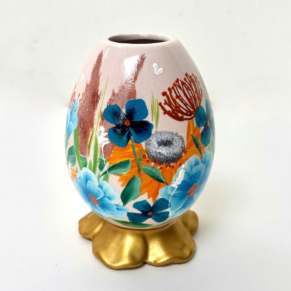 Een eivormige keramische vaas, beschilderd met blauwe en oranje bloemen, rustend op een goudkleurige, bloemvormige sokkel.