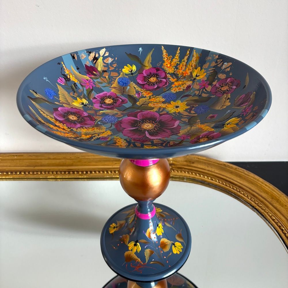 Decoratieve blauwe schaal op voet met kleurrijk, handgeschilderd bloemenmotief en een gouden accent, op een plank.