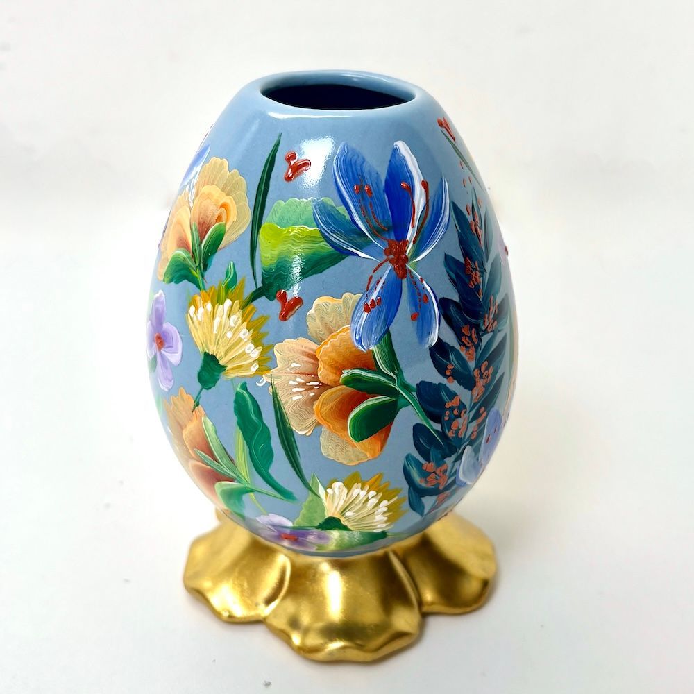Een  lichtblauwe decoratieve vaas, beschilderd met levendige bloemen en groen, rustend op een gouden, bloemvormige voet.