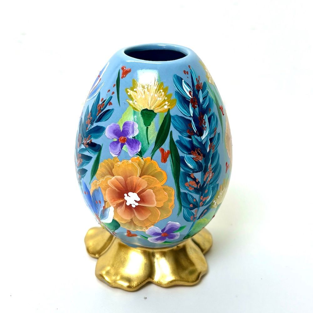 Een  lichtblauwe decoratieve vaas, beschilderd met levendige bloemen en groen, rustend op een gouden, bloemvormige voet.