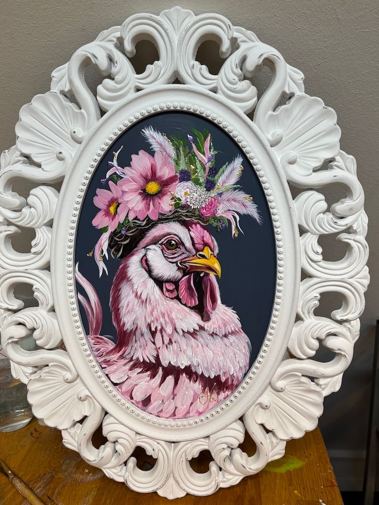 Een kruissteekportret van een pluizig roze kippetje met een bloemenkroon, in een sierlijke witte ovale lijst.