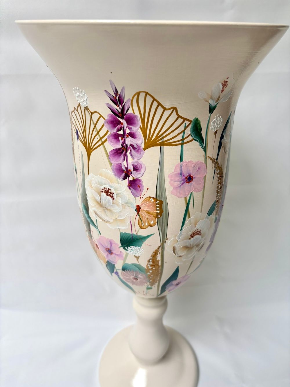Een crèmekleurige vaas op een voetstuk, beschilderd met paarse bloemen, groene bladeren en gouden vlinder- en bloemaccenten.