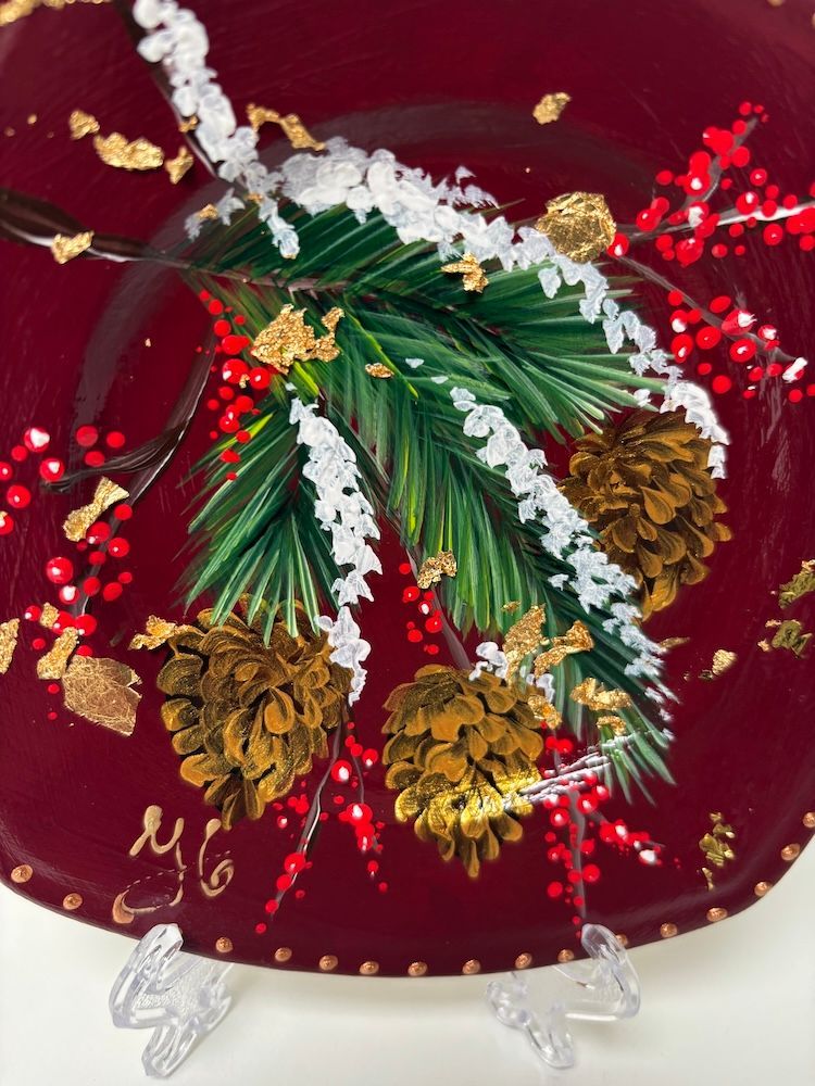 Decoratief rood vierkant bord met groene hulstbladeren en gouden accenten op een transparante standaard.