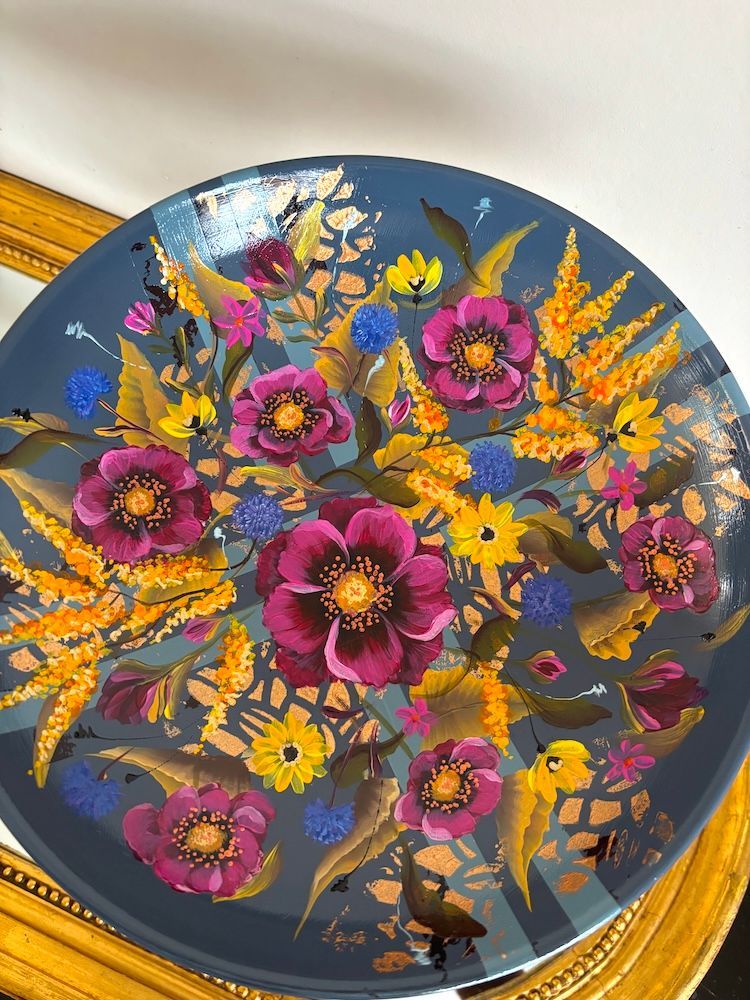 Decoratieve blauwe schaal op voet met kleurrijk, handgeschilderd bloemenmotief en een gouden accent, op een plank.