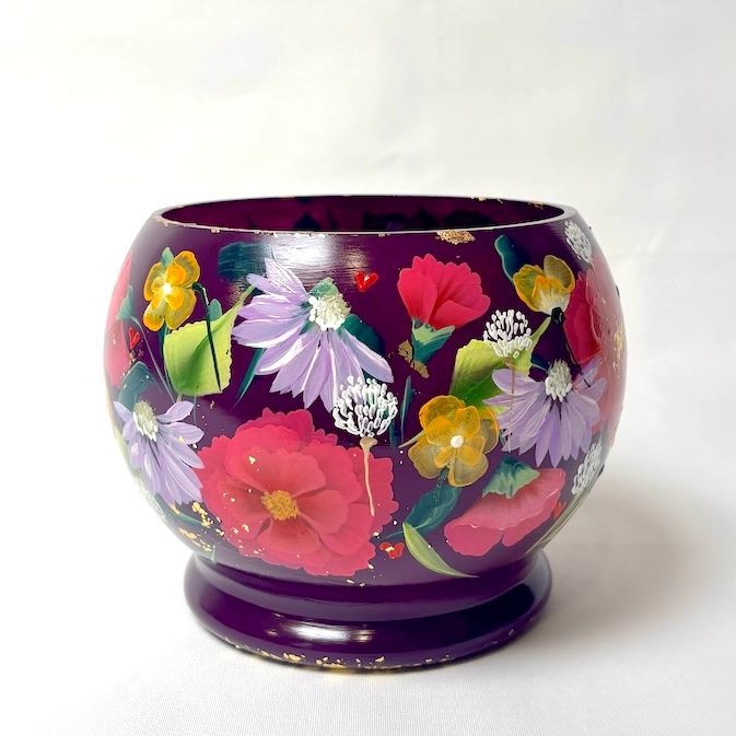 Een donkerpaarse, bolvormige houten schaal met handgeschilderde kleurrijke bloemen, rustend op een voetstuk.