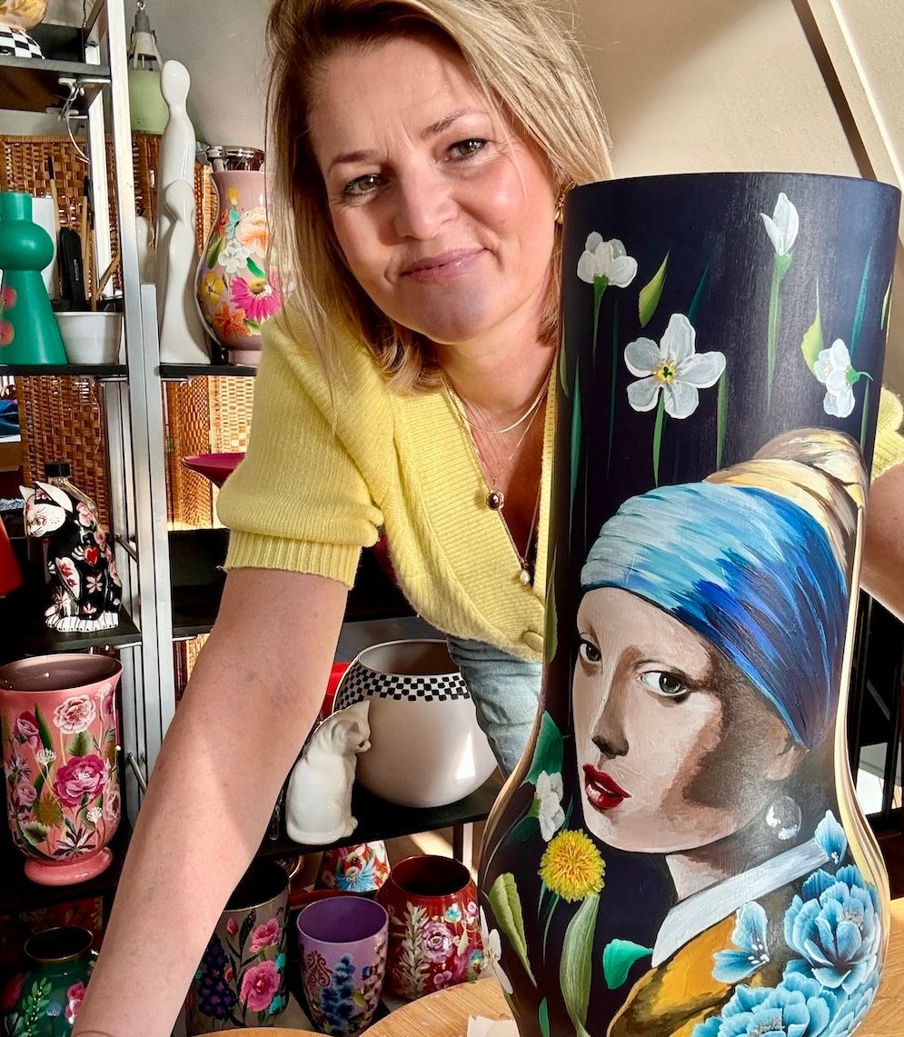 Een kunstenares in een gele top houdt een vaas vast, handbeschilderd met een portret geïnspireerd op het meisje met de parel.